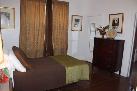 A bedroom with a bed covered in brown and green bedding, two framed pictures on the walls, a dark wooden dresser with decorative items on top, and brown patterned curtains covering a window.
