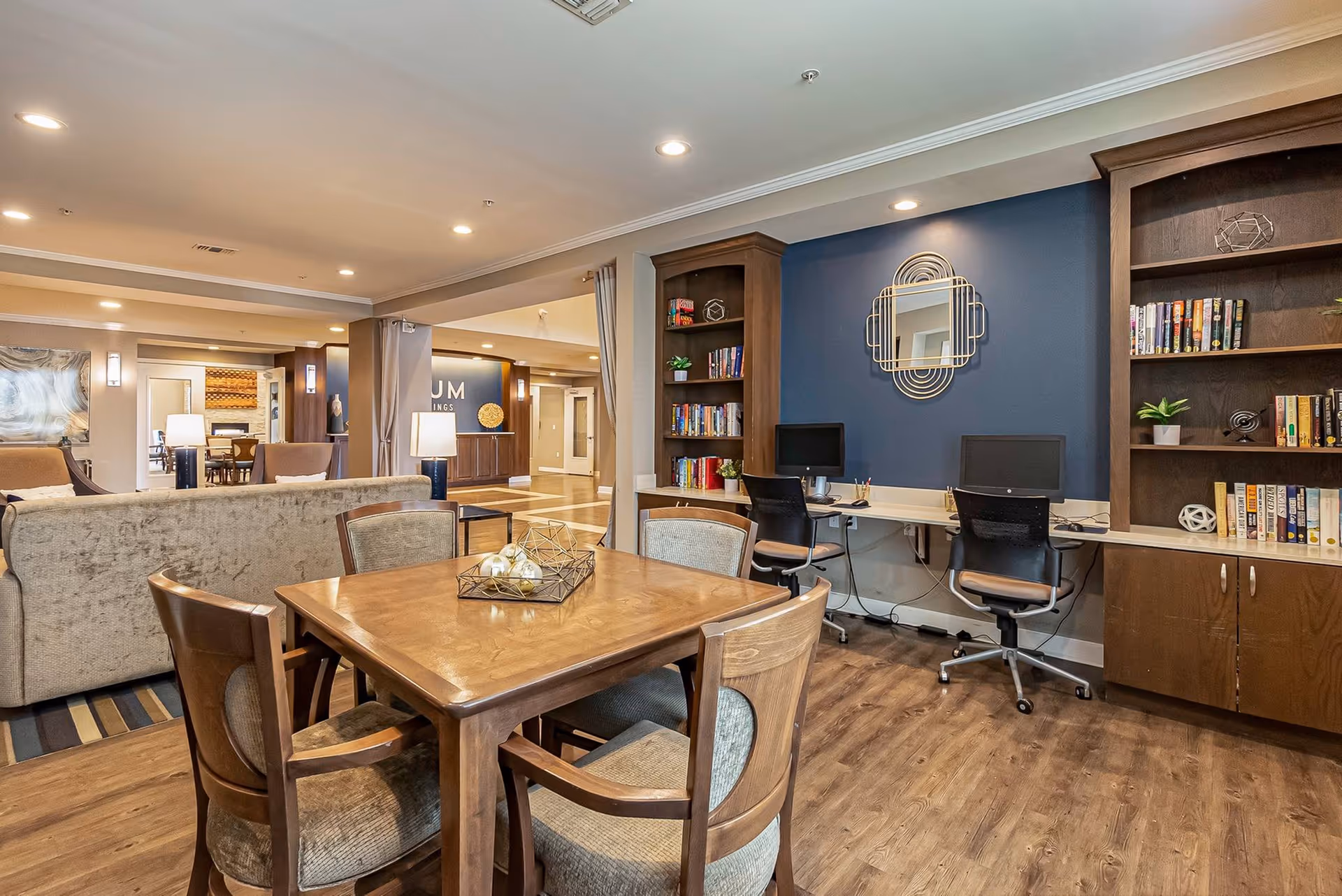 Spacious communal living area with a wooden table and chairs in the foreground, sofas to the left, and a computer workstation with bookshelves against a blue accent wall.
