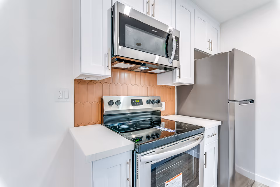 Modern kitchen area featuring a stainless steel electric stove with a glass cooktop, a stainless steel microwave mounted above it, white cabinetry, a beige tile backsplash with a scalloped design, and a stainless steel refrigerator.
