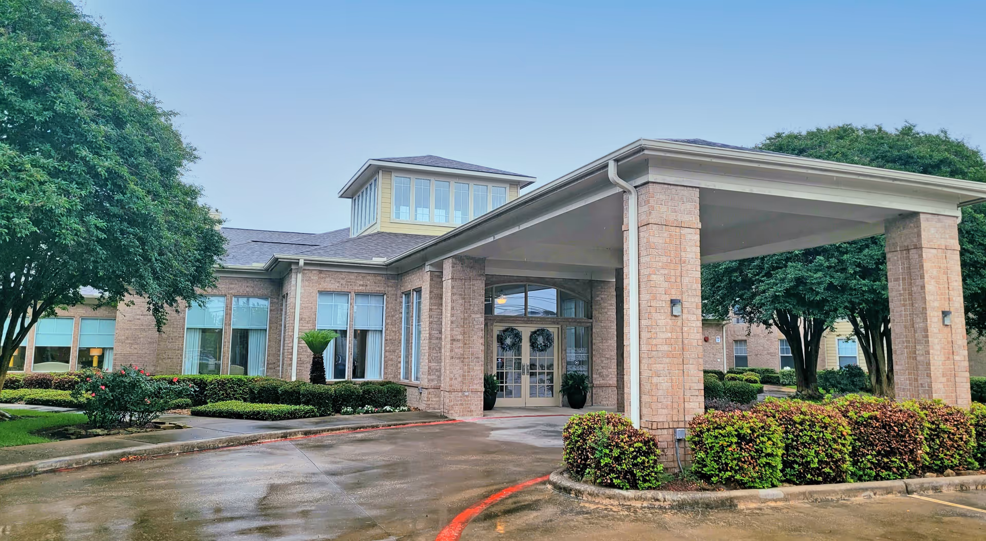 Brick senior living building entrance with a covered porte-cochere, glass doors, trimmed shrubs and a wet driveway.