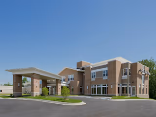 Brick senior living facility with a covered entrance and parking lot under clear skies.