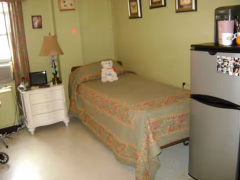 Cozy bedroom with a single bed, teddy bear, nightstand, lamp, and mini-fridge.