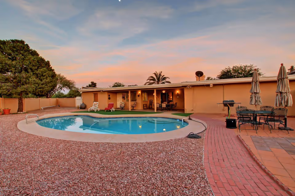 Backyard pool and patio area with lounge chairs, dining set, umbrellas, and a single-story building at sunset.