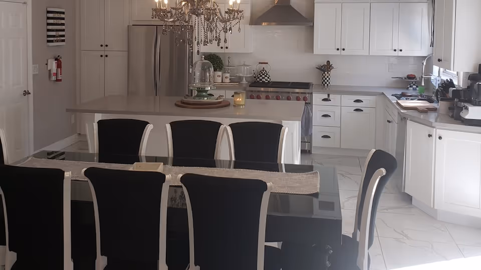 Modern kitchen and dining area with a black glass dining table surrounded by eight black and white chairs. The kitchen features white cabinets, a large island with a chandelier above, stainless steel refrigerator, stove with a hood, and various kitchen appliances on the countertops. The floor is tiled with white marble-like tiles.