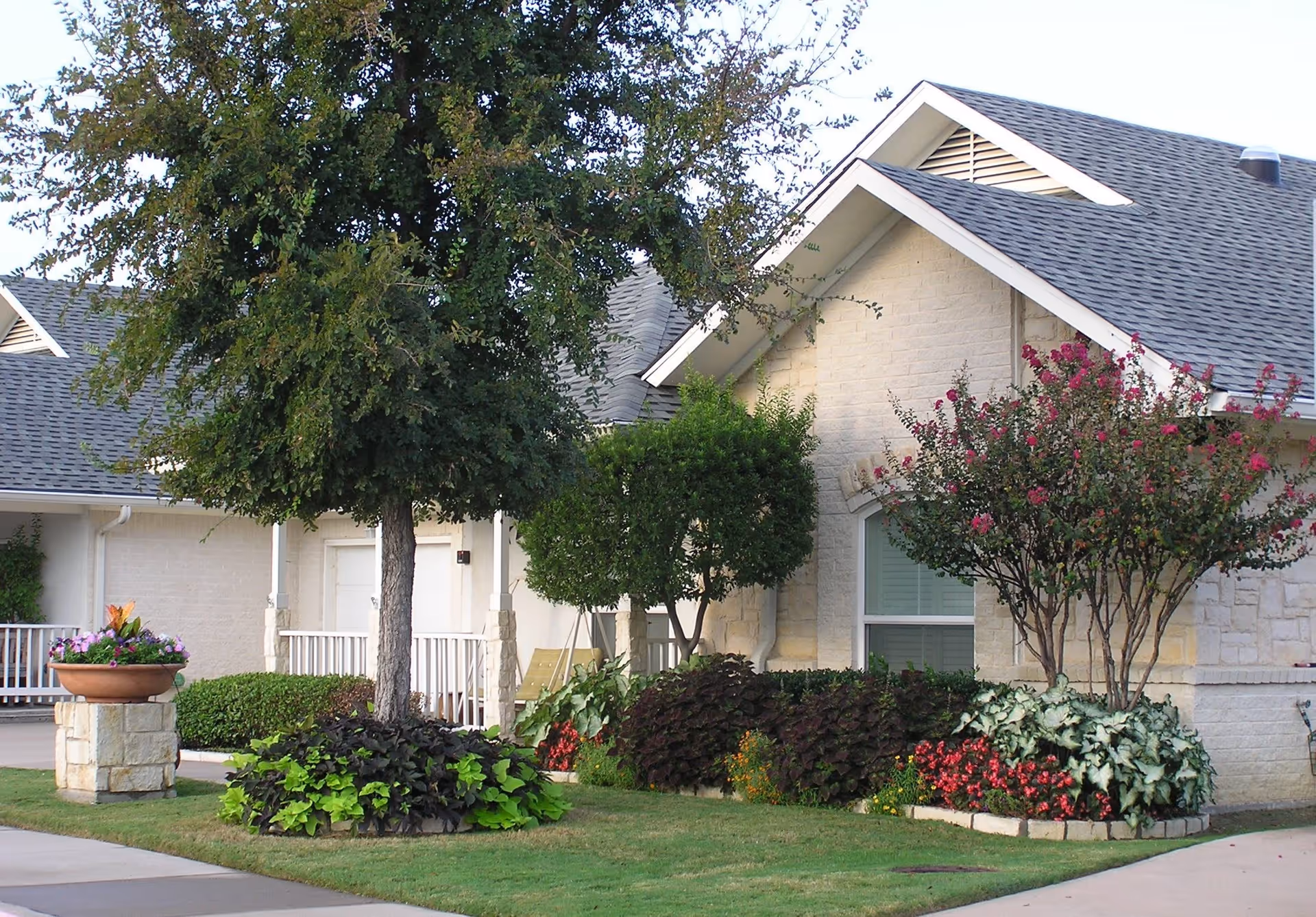 Front of a single-story stone building with a pitched roof, porch, trees, and landscaped flower beds.
