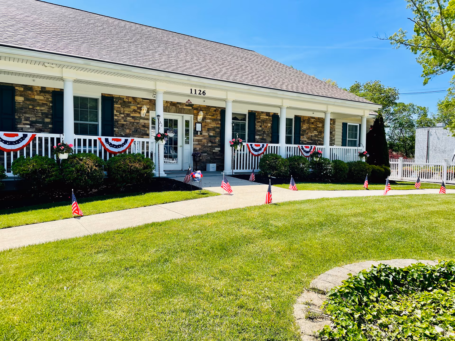 Front exterior view of a single-story building with stone and brick facade, white columns, and a porch decorated with red, white, and blue bunting and small American flags lining the walkway. The building number 1126 is visible above the entrance door. The lawn is green and well-maintained with a circular garden bed in the foreground.