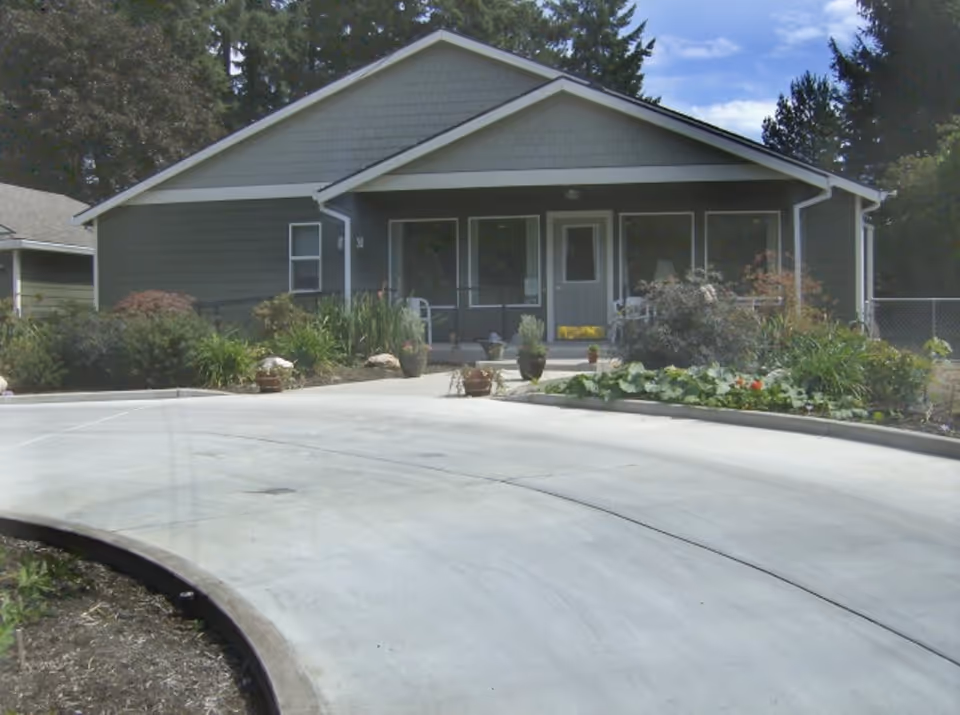 Front exterior view of a single-story building with a covered porch, surrounded by plants and shrubs, with a curved concrete driveway in front.