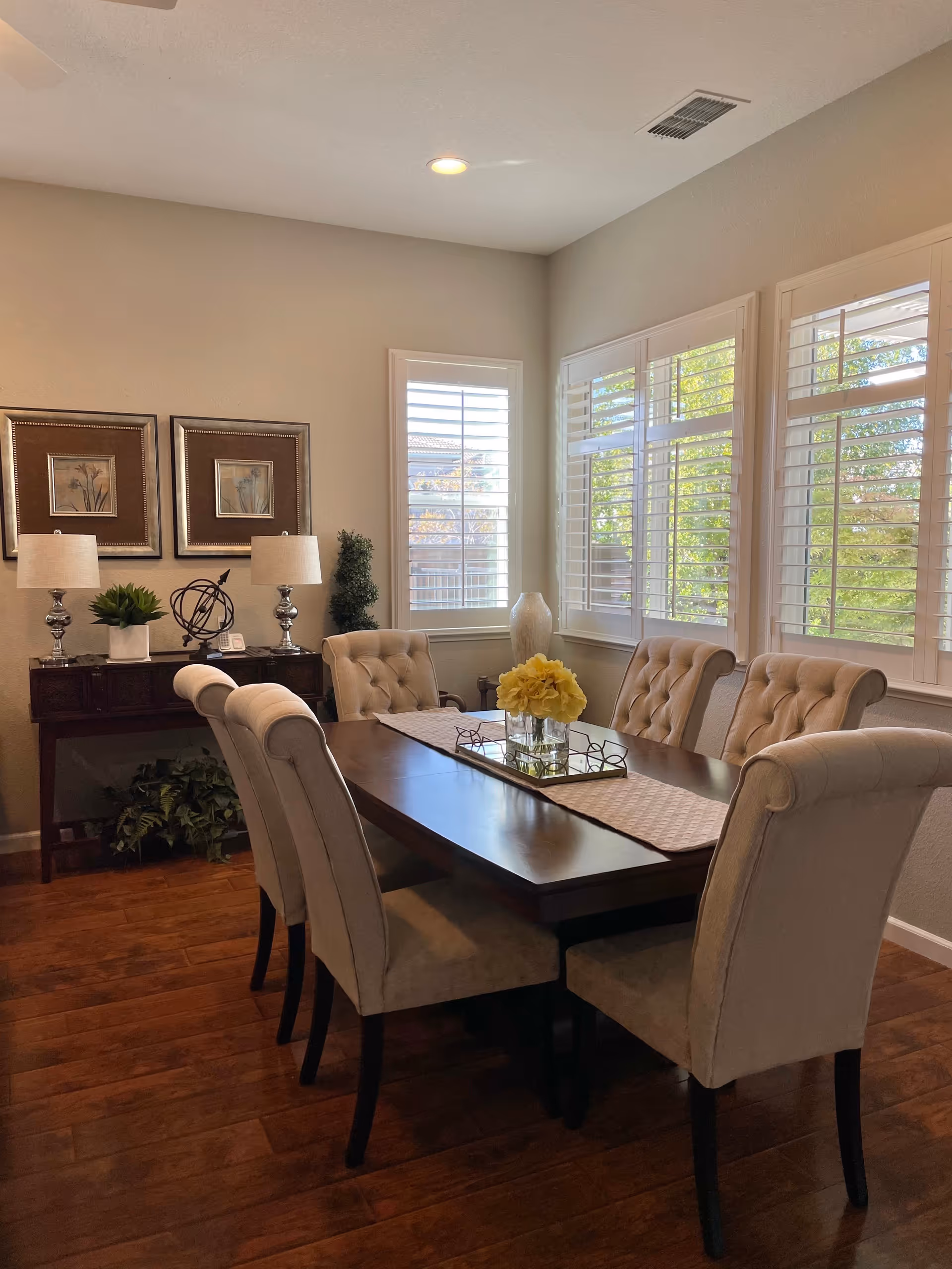 A dining room with a dark wooden table surrounded by six beige upholstered chairs. The table has a beige runner with a glass vase of yellow flowers and a decorative tray. The room has wooden flooring, three large windows with white plantation shutters letting in natural light, and a sideboard with two lamps, framed artwork, and decorative plants.