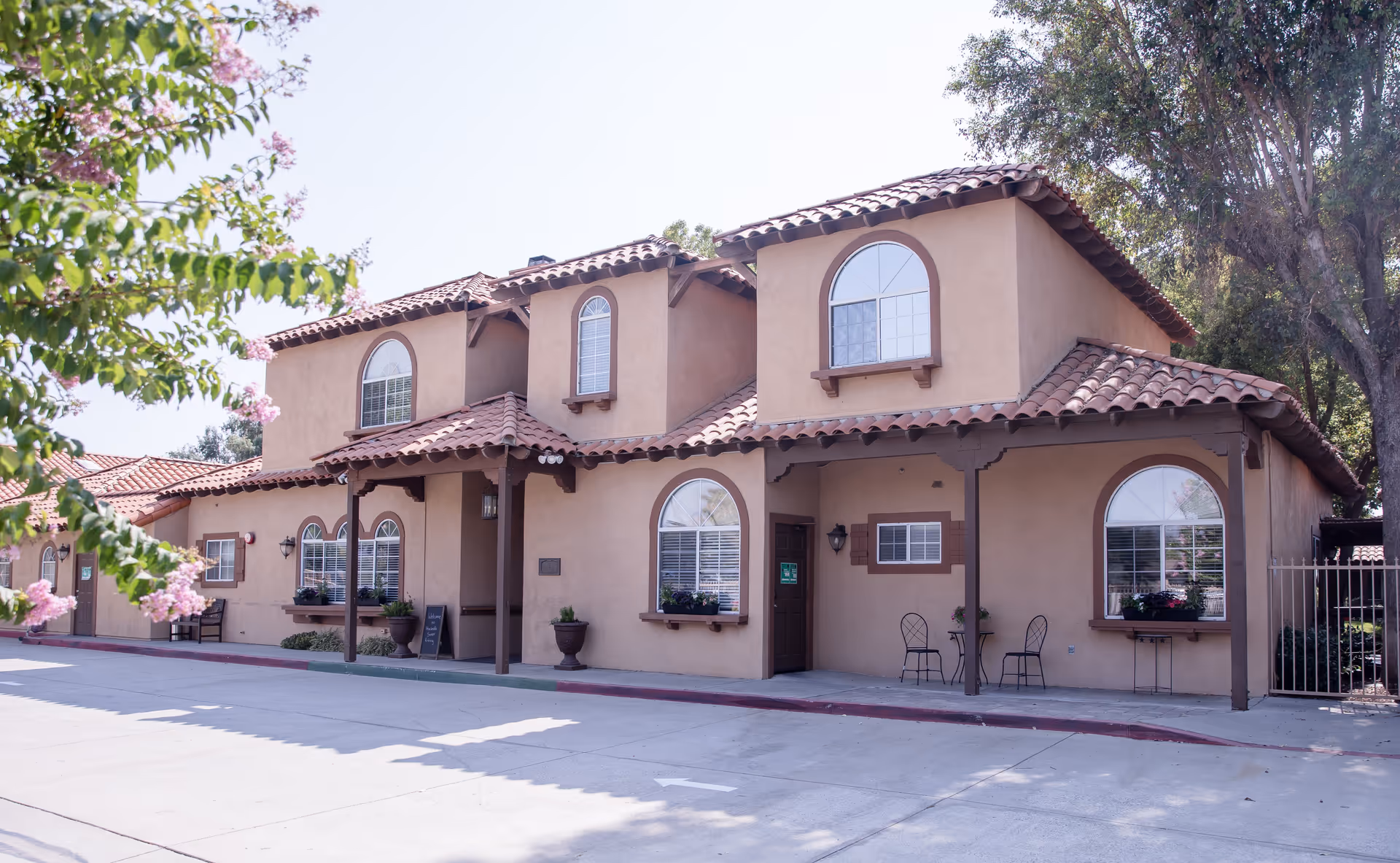 Exterior view of a two-story building with beige stucco walls and red tile roof, featuring arched windows with white shutters and flower boxes. There is a covered porch area with two chairs and a small table, surrounded by trees and flowering plants.