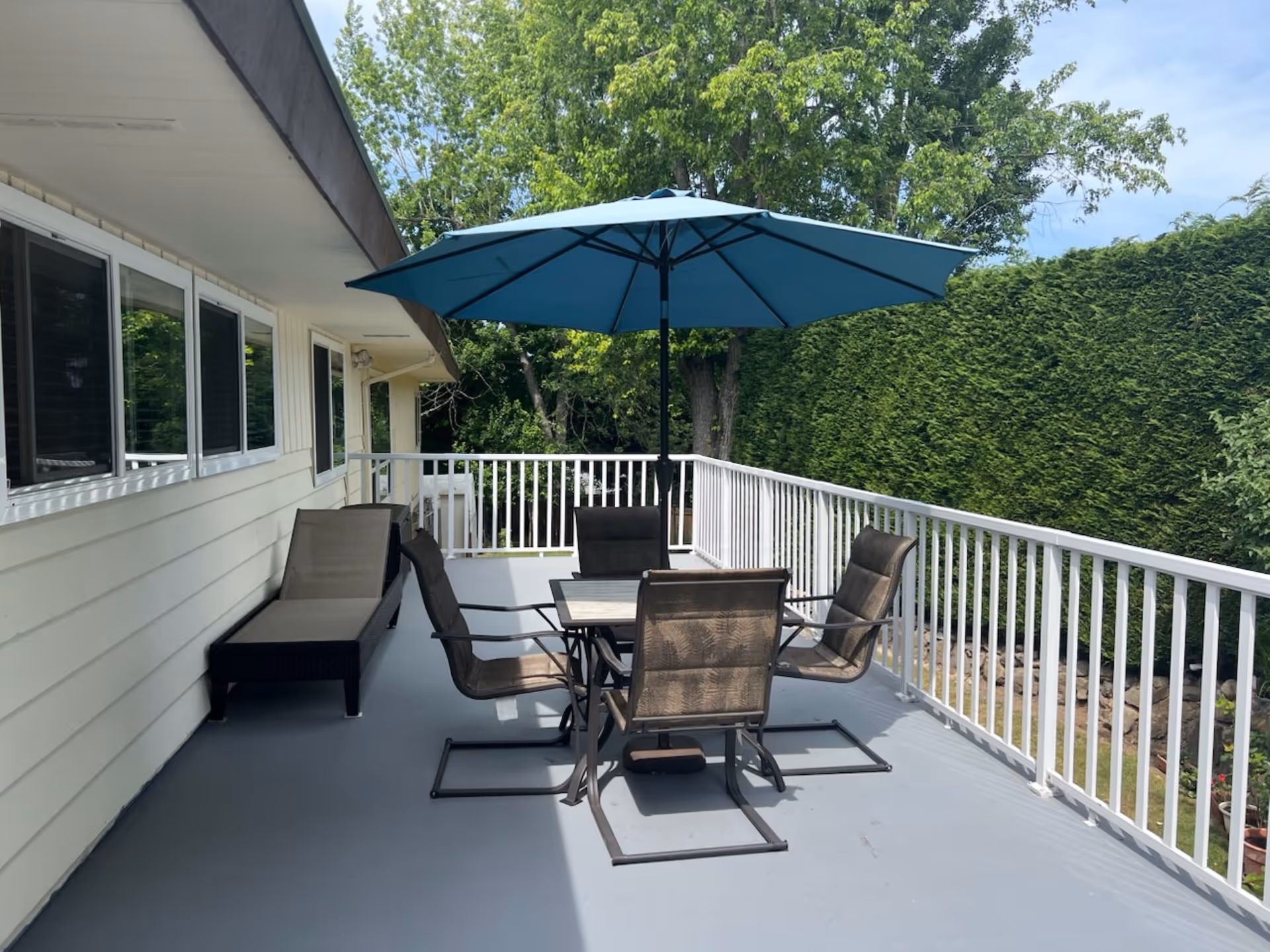 Outdoor deck with a table, several chairs and a blue umbrella next to a white railing and hedges.