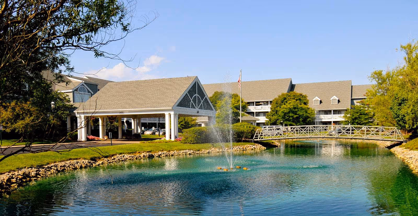 A serene outdoor scene at StoryPoint Cincinnati featuring a pond with a water fountain in the center, a small arched pedestrian bridge over the pond, and a large building with a covered entrance and multiple windows in the background. Trees and greenery surround the area under a clear blue sky.
