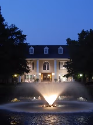 Evening view of a large building with lit windows and a central entrance, framed by trees on both sides. In the foreground, a water fountain is illuminated, spraying water upwards in front of the building.
