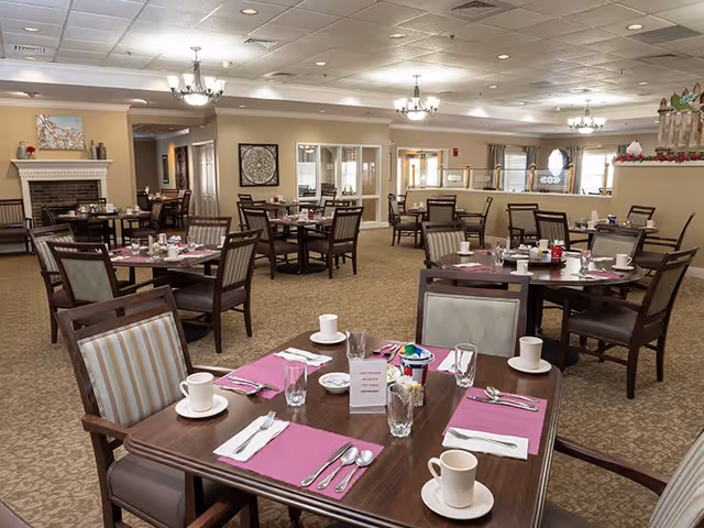 A spacious dining room with multiple wooden tables and chairs arranged neatly. Each table is set with pink placemats, white cups, glasses, and silverware. The room has beige carpet, cream-colored walls, and a white ceiling with recessed lighting and chandeliers. There is a fireplace on the left side and windows along the back wall allowing natural light to enter.