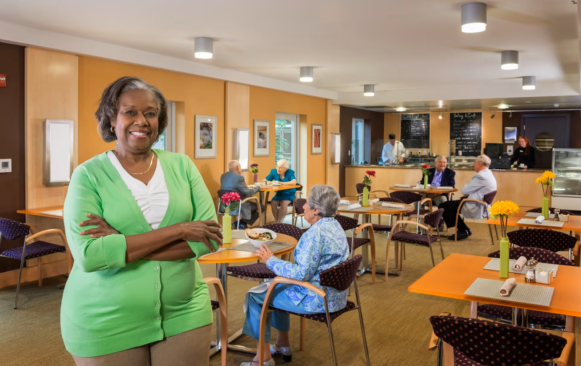 A smiling woman in a green cardigan stands with her arms crossed in a dining room of a retirement community. Behind her, several elderly people are seated at tables eating and conversing. The room has warm lighting, wooden tables with flowers in vases, and a counter area with staff serving food.