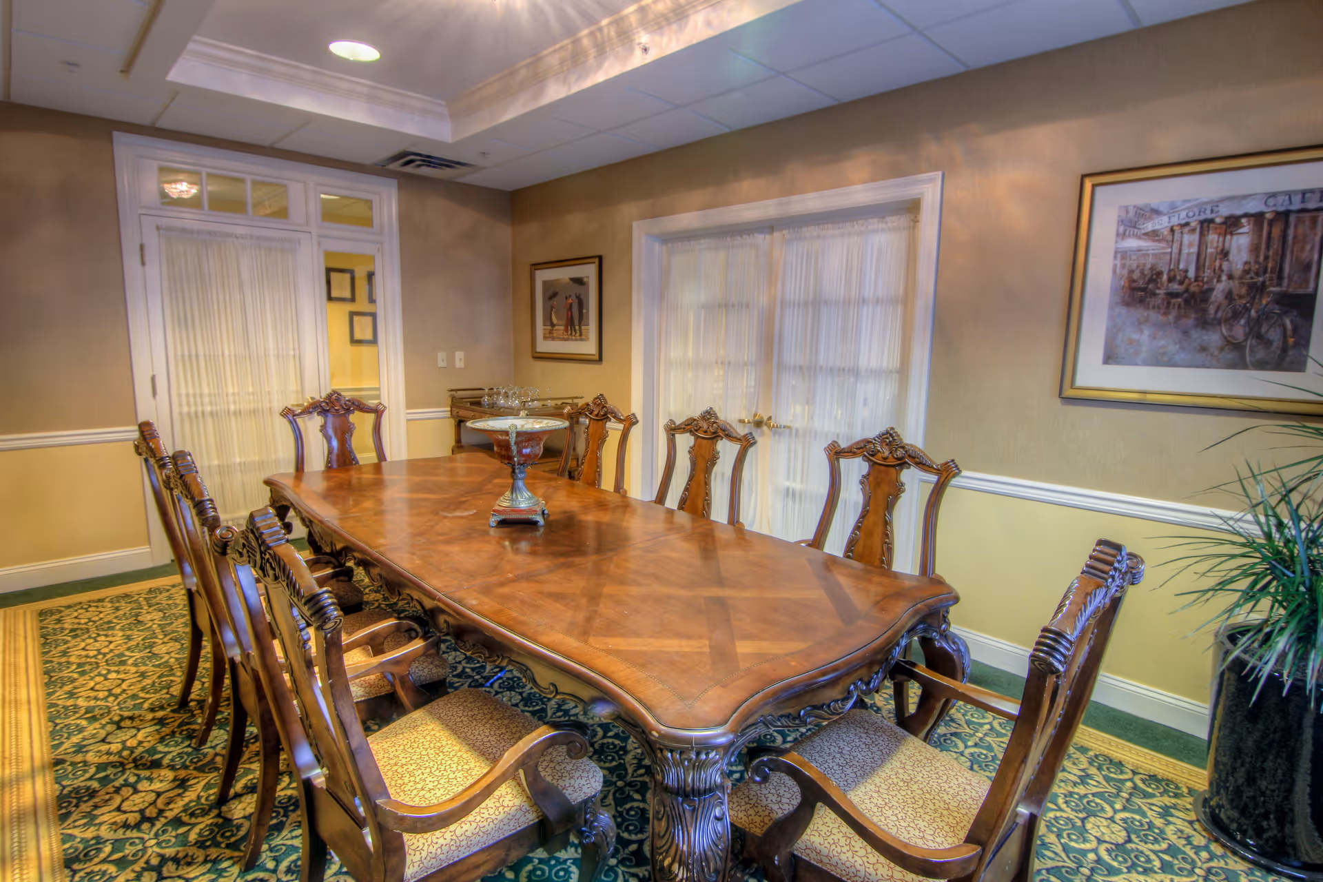 A formal dining room with a long wooden table surrounded by eight ornate wooden chairs with upholstered seats. The room has beige walls with white trim, a patterned carpet, framed artwork on the walls, and a large window with sheer curtains. There is a decorative centerpiece on the table and a potted plant in the corner.