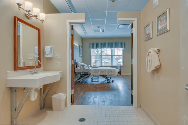 View from a bathroom into a patient bedroom with a hospital bed by a large window and a sink in the foreground.