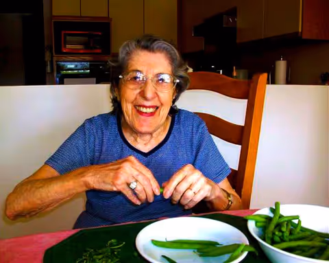 An elderly woman with glasses and short gray hair is sitting at a table in a kitchen, smiling while preparing green beans. She is wearing a blue striped shirt and is seated on a wooden chair. There are two white bowls on the table, one with green beans and the other with trimmed green beans.