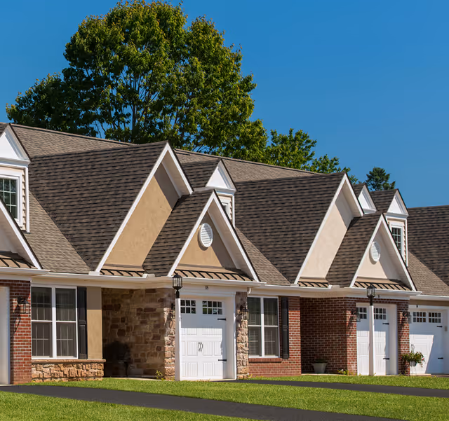 Front exterior of a row of attached residential units with pitched gable roofs, garage doors, brick and stone facades, and a lawn.