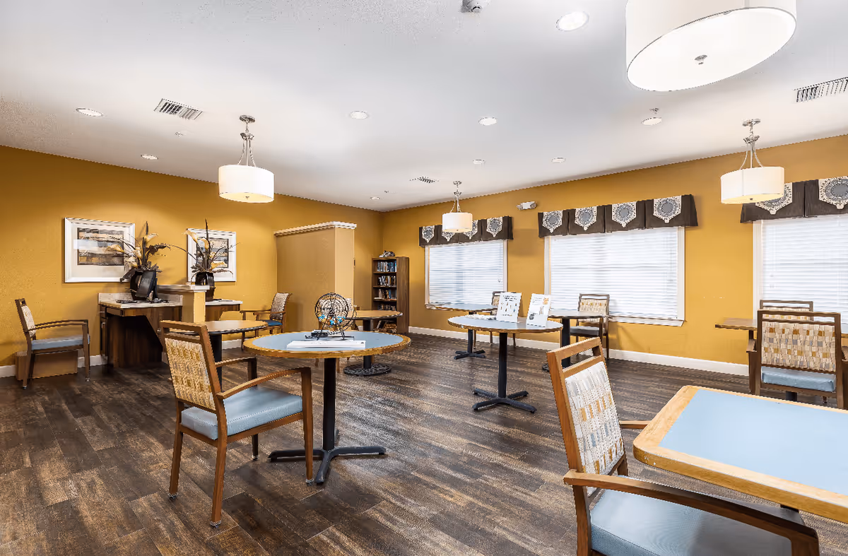 A spacious room with multiple round and square tables surrounded by wooden chairs with cushioned seats and backs. The walls are painted mustard yellow, and there are three large windows with valances letting in natural light. The floor is dark wood, and the ceiling has several recessed lights and hanging pendant lamps. There is a small desk area with a computer and decorative plants in the corner.