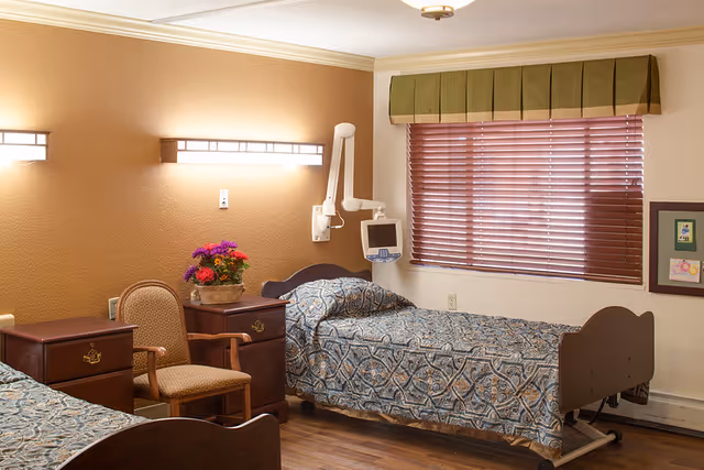 A shared senior bedroom with two twin hospital-style beds, wooden nightstands, a chair, wall-mounted medical equipment and lighting, and a window with blinds.