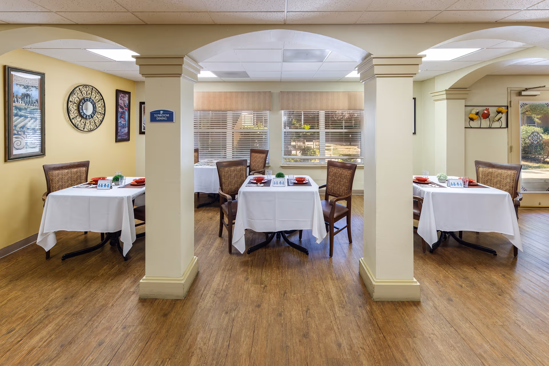 A sunroom dining area with three tables covered in white tablecloths, each set with red plates and glasses. The room has wooden flooring, cream-colored walls, decorative wall art, and large windows letting in natural light. There are three archways supported by columns dividing the space.