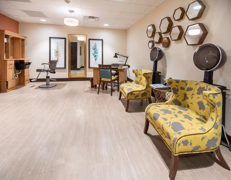 A bright and clean salon area in a senior living facility featuring two yellow floral-patterned chairs with hair dryers behind them, a wooden styling station with a salon chair, a desk with two chairs, a large mirror, and two framed botanical prints on the wall.