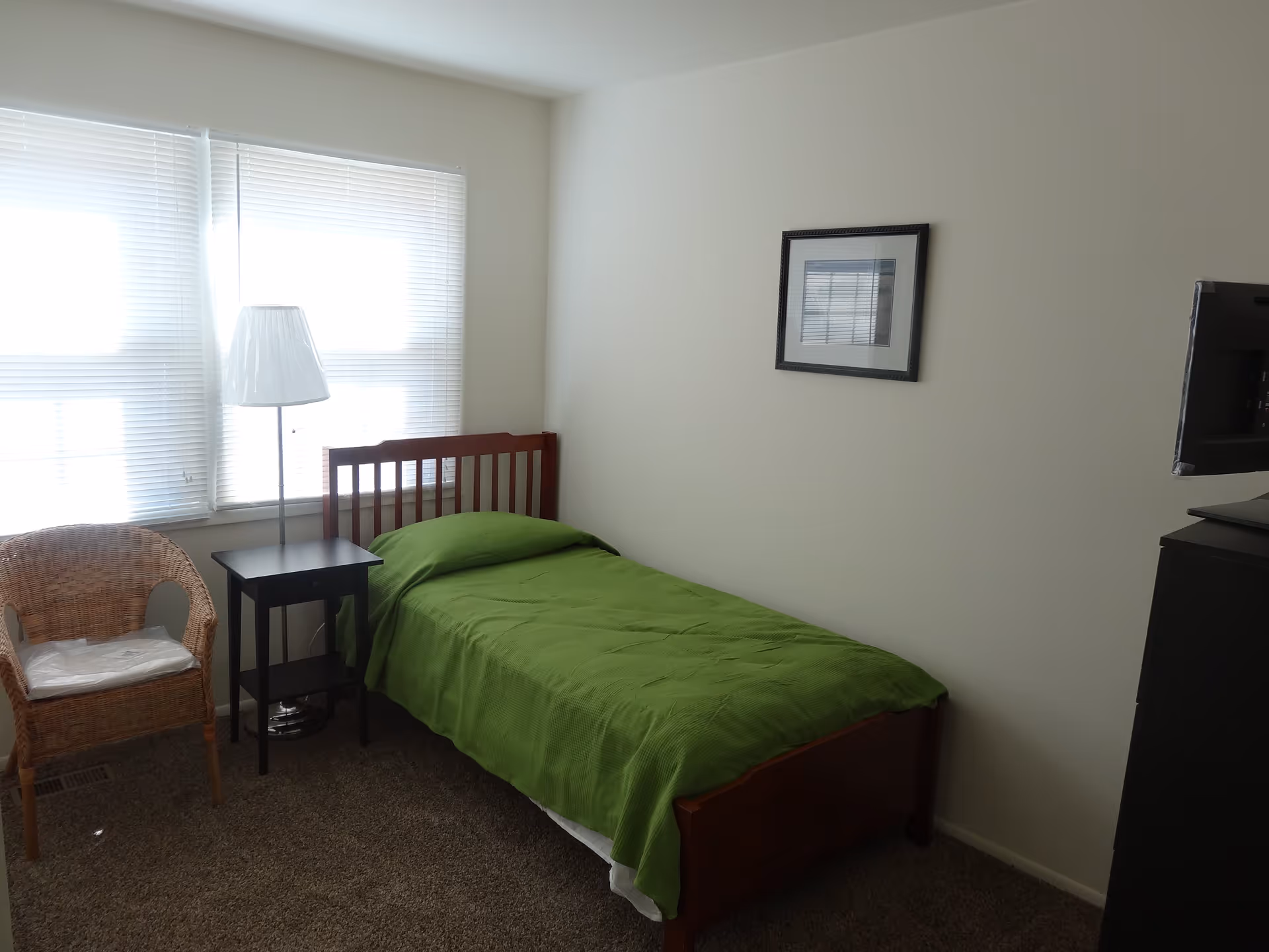 A small bedroom with a single wooden bed covered with a green blanket. Next to the bed is a black nightstand with a white floor lamp. There is a wicker chair with a cushion near the window with white blinds. A framed picture hangs on the plain white wall, and a TV is partially visible on the right side.