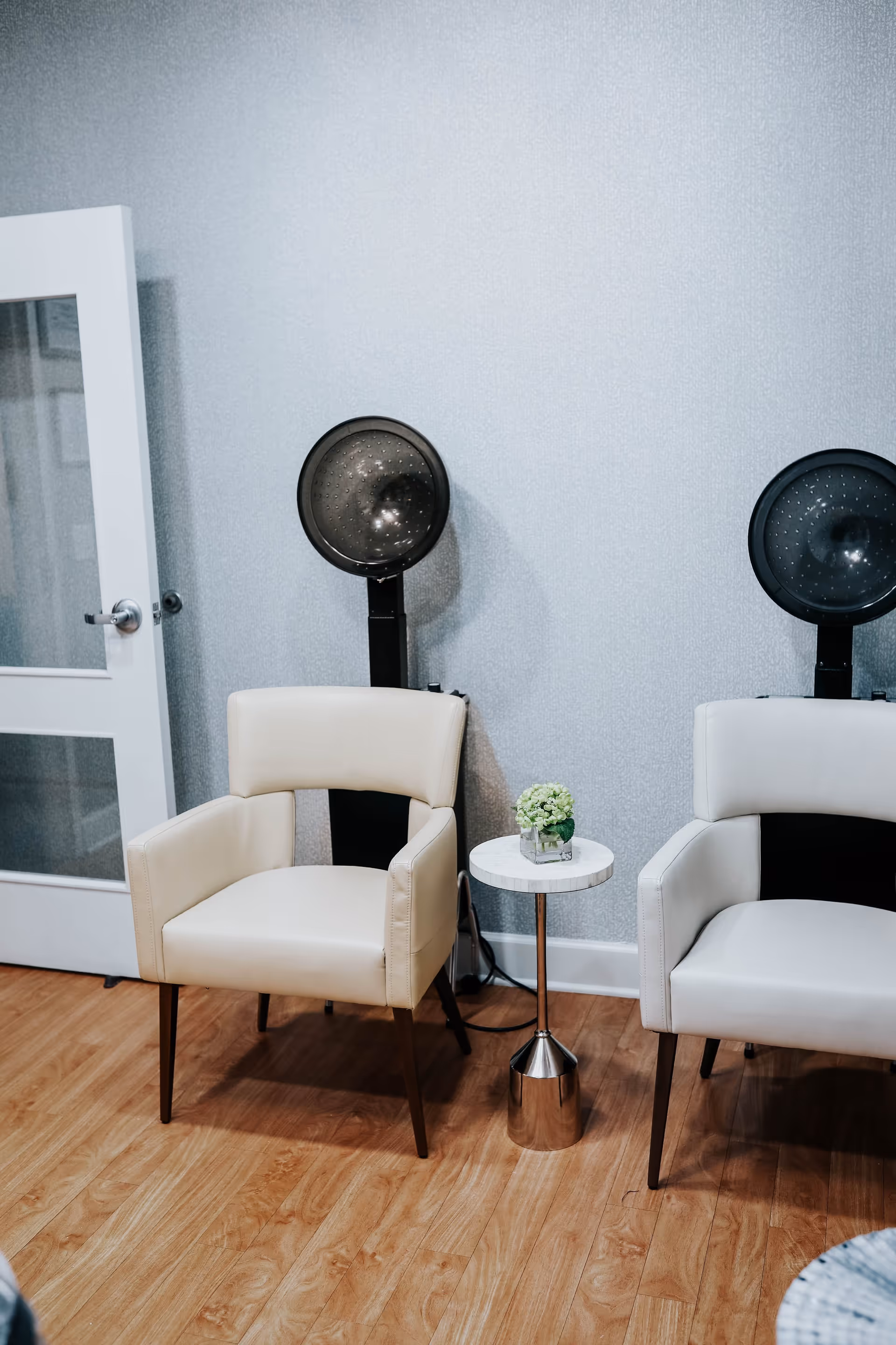 Two cream salon chairs under hooded hair dryers with a small round side table holding a plant in a light, wood-floored room.