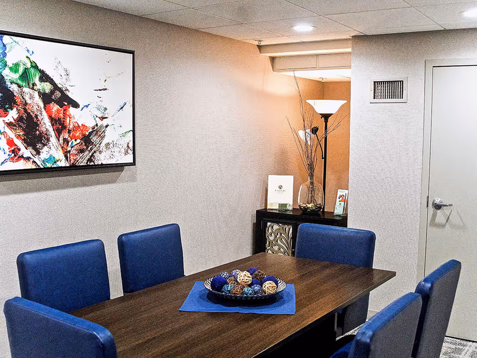 A small meeting or dining area with a rectangular wooden table surrounded by six blue upholstered chairs. On the table is a decorative bowl filled with assorted decorative balls on a blue placemat. The room has beige walls, a white door, a floor lamp, a small cabinet with decorative items, and a colorful abstract painting on the wall.