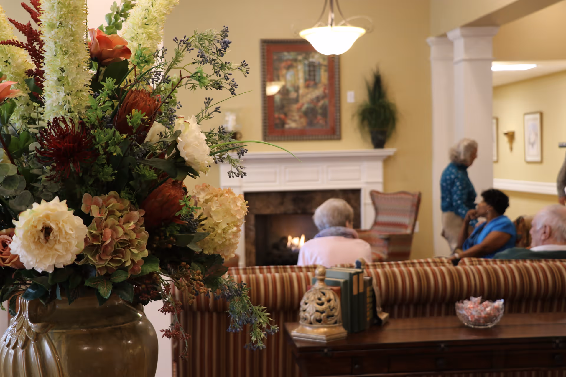 A cozy living room area in a senior living facility with a large floral arrangement in the foreground. In the background, several elderly people are seated and conversing near a lit fireplace, with framed artwork and a potted plant on the wall.