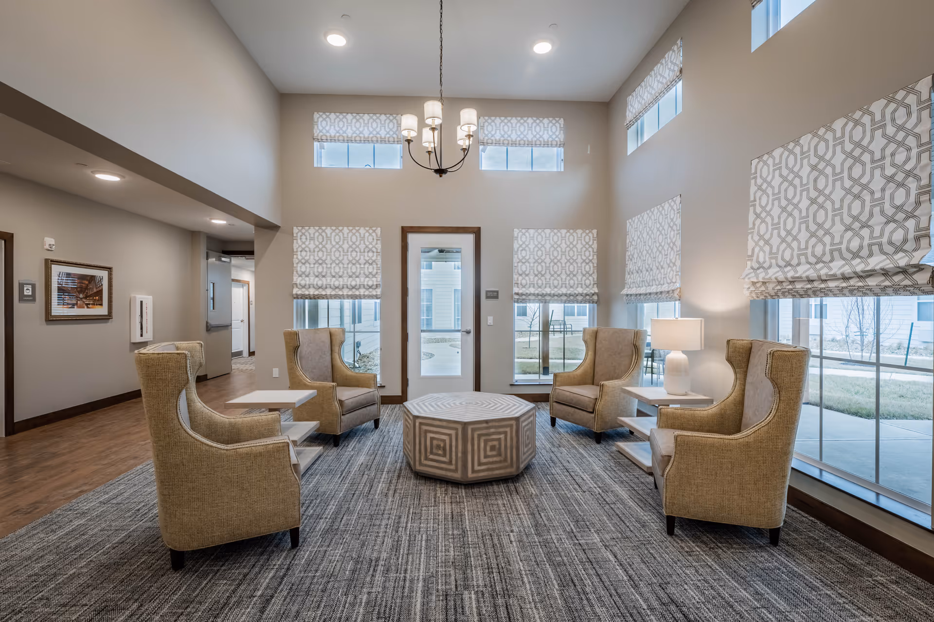 A bright and spacious senior living facility common area with four beige upholstered armchairs arranged around a geometric patterned octagonal ottoman on a gray carpet. The room features large windows with patterned Roman shades, a white table lamp on a side table, and a modern chandelier hanging from the high ceiling. The walls are painted light beige, and there is a door leading outside with windows on either side.