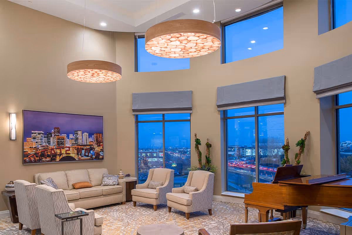 A spacious and well-lit living room with large windows showing a cityscape at dusk. The room features a beige sofa, two armchairs, a grand piano, and modern circular ceiling lights. There is a large framed cityscape artwork on the wall and decorative plants near the windows.
