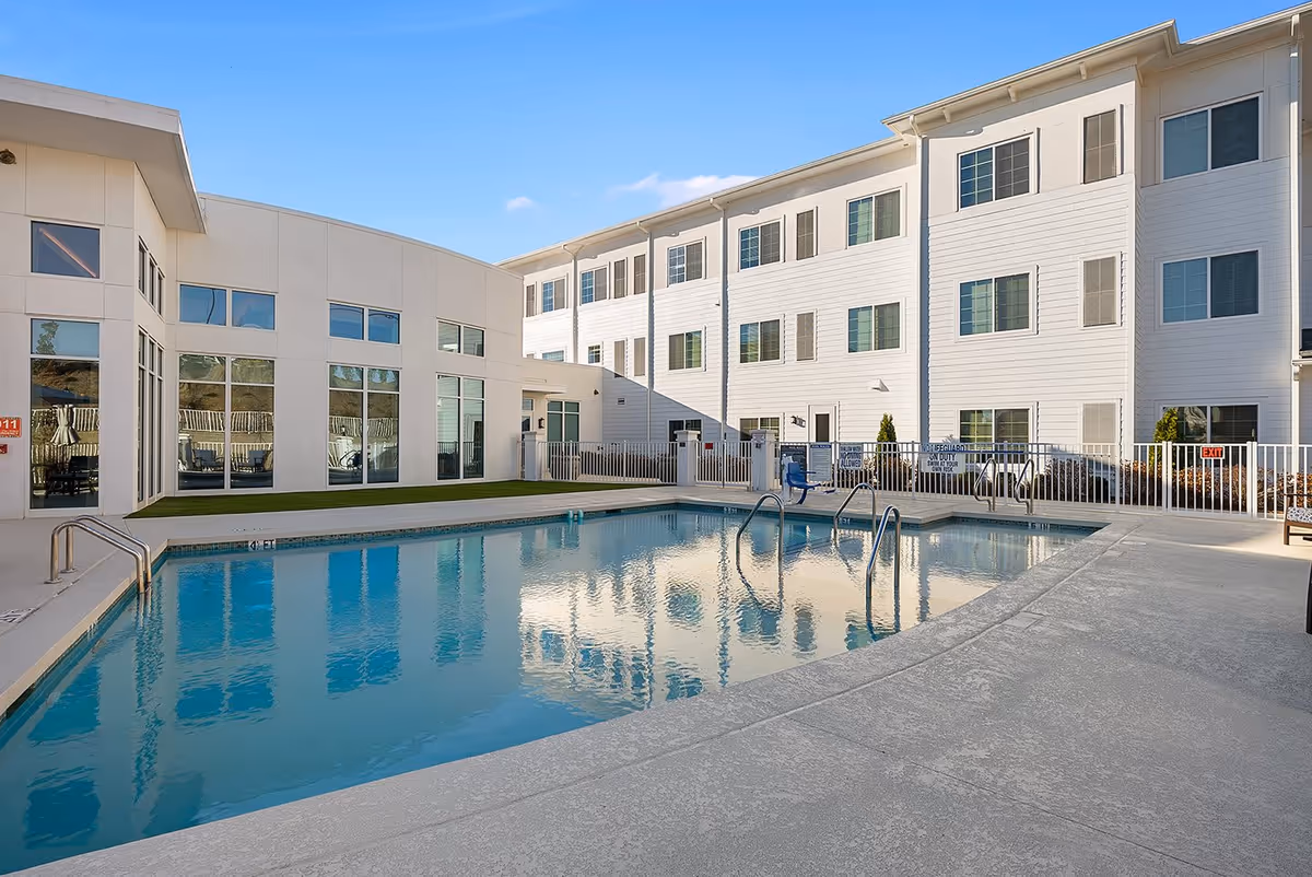 Outdoor swimming pool area at The Meridian at Carolina Lakes with clear blue water, surrounded by a concrete deck and a white fence. The pool is adjacent to a modern three-story building with multiple windows reflecting in the water. The sky is clear and blue.
