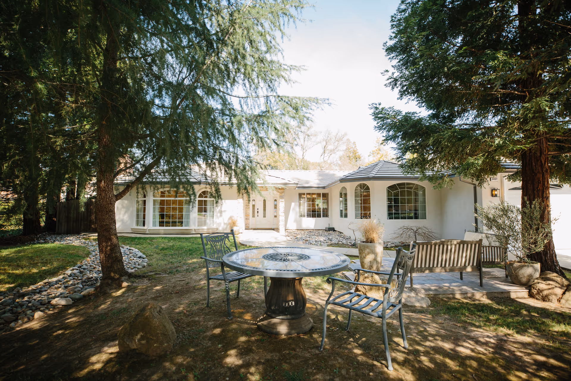 Single-story house front with large windows, trees, and an outdoor seating area with a round table and chairs.