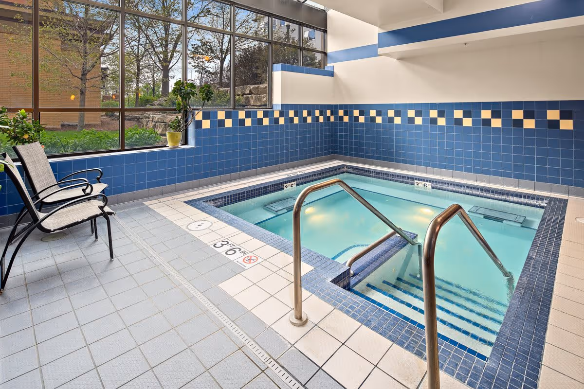 Indoor hot tub with blue and white tiled walls and floor, metal handrails leading into the water, two chairs on the left side, and large windows showing trees and outdoor greenery.