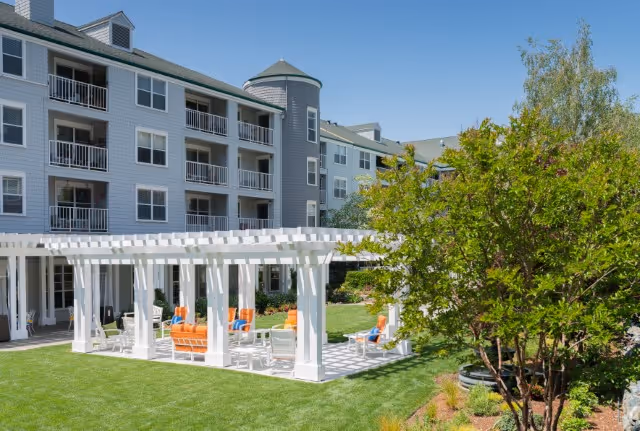 Outdoor seating area with white pergola and orange cushioned chairs on a green lawn in front of a multi-story senior living facility building under a clear blue sky.