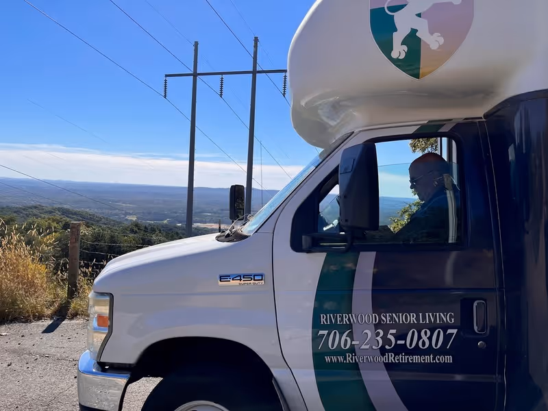 A Riverwood Senior Living shuttle van parked at a scenic overlook with a driver visible inside and rolling hills in the background.