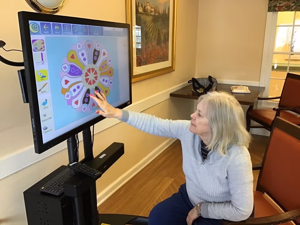 An elderly woman seated in a chair interacts with a large touchscreen monitor mounted on a stand, displaying a colorful digital drawing or painting application. The room has wooden flooring, beige walls, framed artwork, and red cushioned chairs.