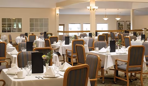 A spacious dining room with multiple tables covered in white tablecloths, each set with napkins, cups, glasses, and black menus. The room features cushioned wooden chairs, soft lighting from ceiling fixtures, and large windows allowing natural light.