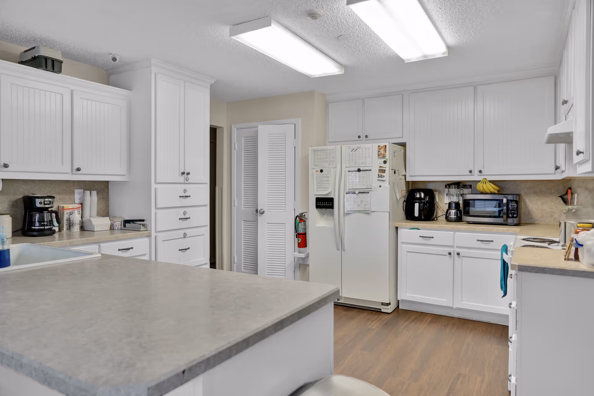 Communal kitchen featuring white cabinets, a central island, refrigerator covered with notes, countertop appliances, and wood-look flooring.