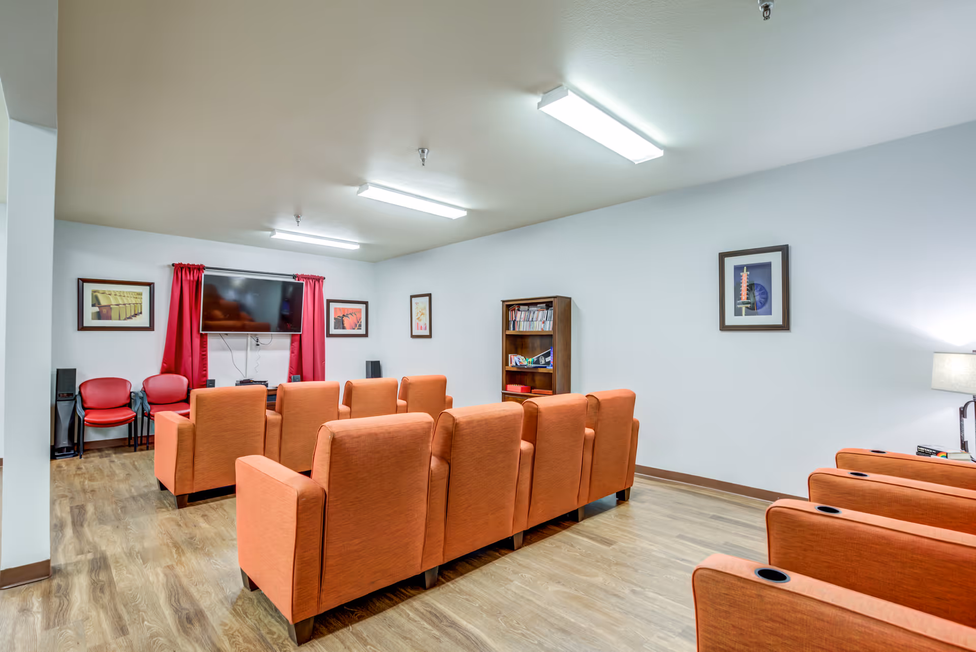 Small media room with rows of orange upholstered theater-style chairs facing a wall-mounted TV, red curtains, framed art, and a bookshelf.