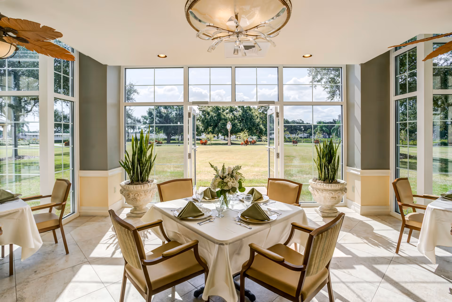 A bright dining room with large floor-to-ceiling windows and glass doors opening to a green outdoor area. The room features a square table set for four with beige chairs, white tablecloth, green folded napkins, glassware, and a floral centerpiece. Two large potted plants flank the open doors, and a decorative ceiling light fixture is visible.