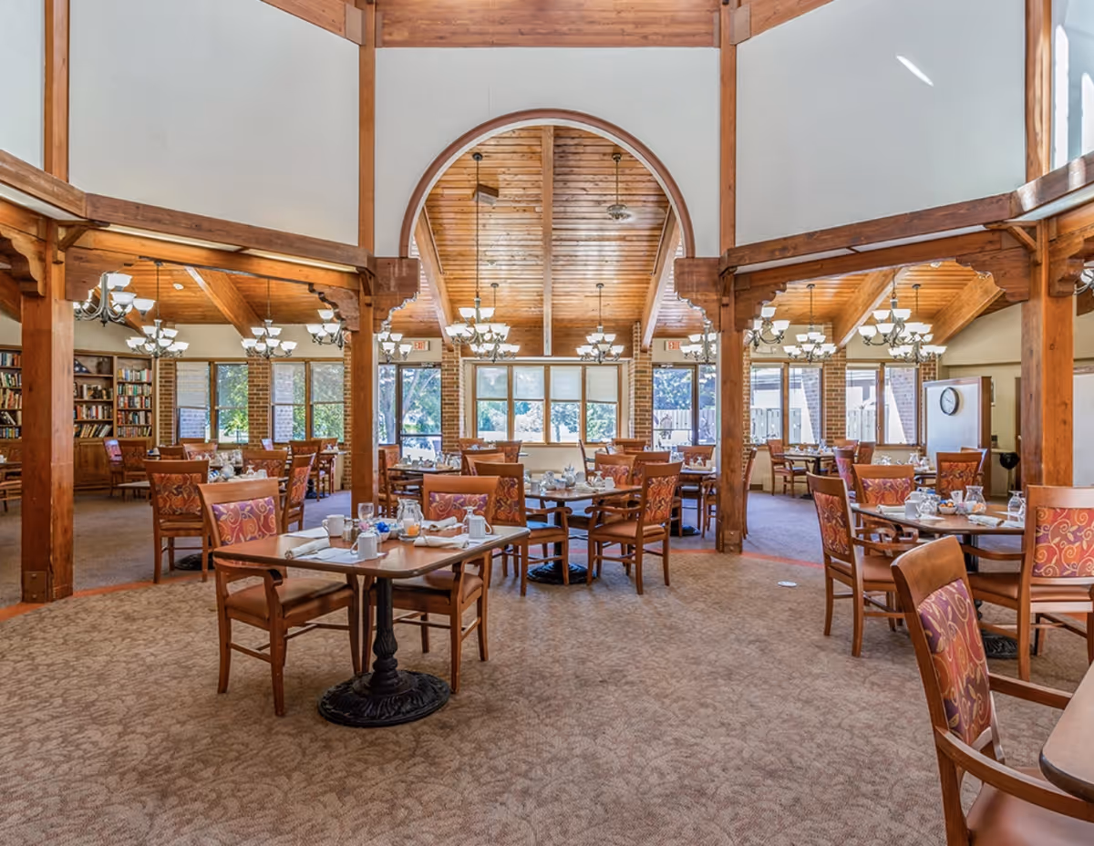 Spacious dining room with wooden beams and vaulted ceiling, tables and chairs set for meals under multiple chandeliers.