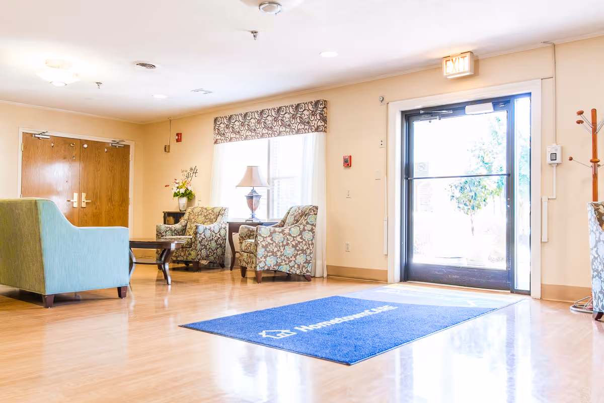 Bright lobby seating area with patterned armchairs, a green sofa, coffee table and a glass entrance door under an EXIT sign.