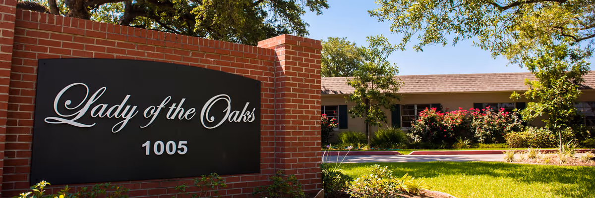 Brick entrance sign reading "Lady of the Oaks" and "1005" in front of a low residential building with trees, shrubs, and a manicured lawn.
