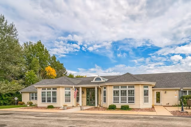 Exterior view of a single-story beige building with multiple large windows, an American flag near the entrance, and a paved driveway in front. The sky is partly cloudy with patches of blue, and there are trees with green and some yellow leaves in the background.