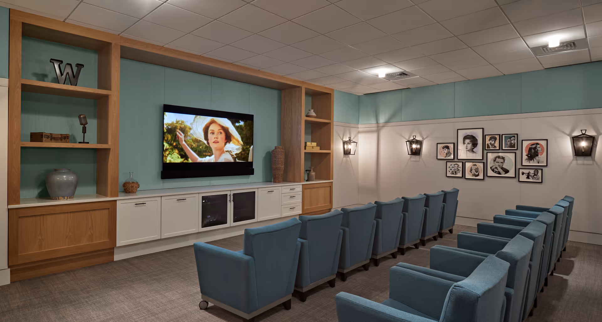 A cozy media room with two rows of blue upholstered chairs facing a wall-mounted flat-screen TV displaying a woman in a hat. The room features built-in wooden shelves and cabinets, soft lighting from wall sconces, and framed black-and-white portraits on the wall.