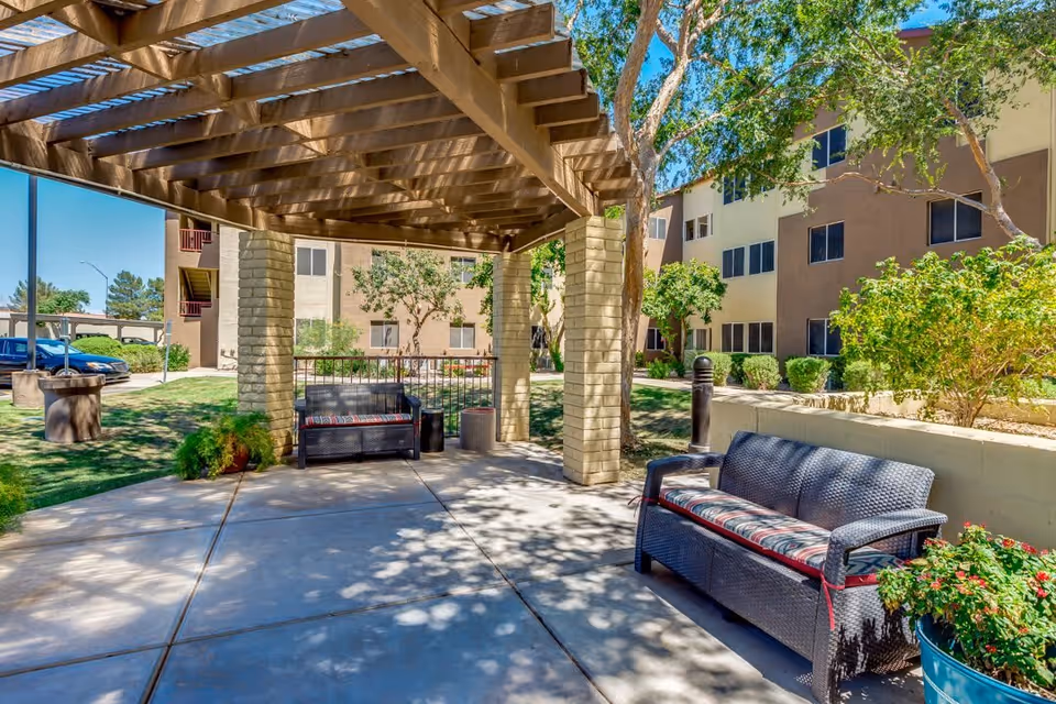 Outdoor seating area at Discovery Point Retirement Community featuring two cushioned wicker benches under a wooden pergola with trees, shrubs, and a multi-story building in the background.