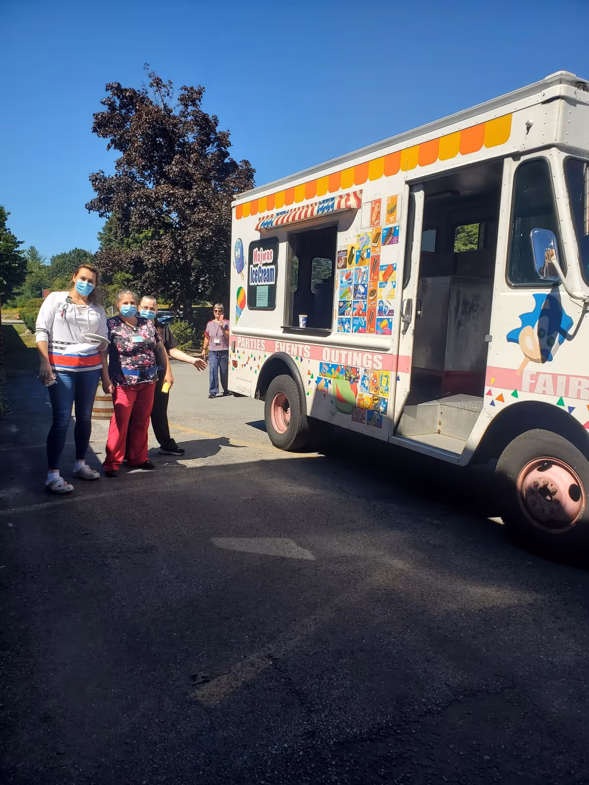 Four people wearing face masks standing outside next to a colorful ice cream truck on a sunny day with trees and clear blue sky in the background.