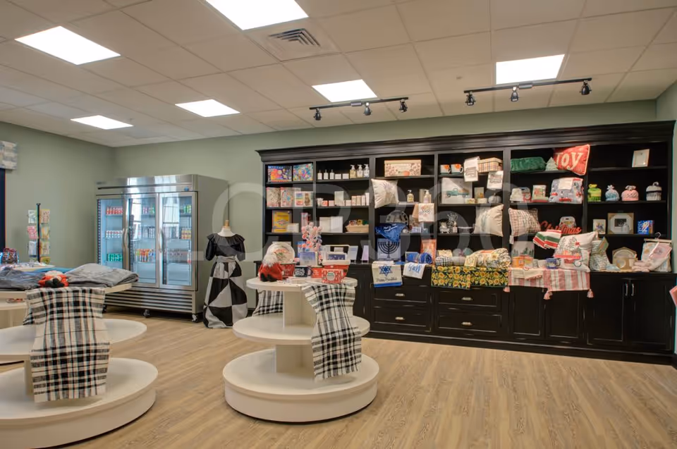 Interior view of a gift shop area in a senior living facility with shelves and round display tables showcasing various gift items, pillows, and decorative pieces. A refrigerated display case is visible in the background.